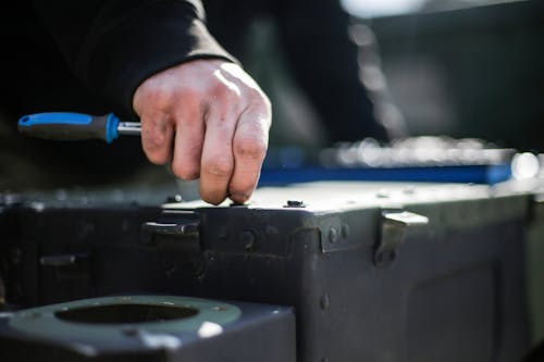 Unrecognizable crop male mechanic in black workwear checking metal details with screwdriver while working outside garage with car