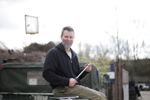 Side view of smiling middle aged man in black hoodie sitting on metal trunk of car outside garage and looking at camera