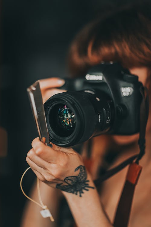 Crop unrecognizable female photographer with tattoo on hand taking pictures on professional photo camera against dark wall