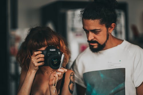 Concentrated female photographer in casual clothes standing near ethnic bearded man and taking pictures on professional photo camera in modern photography school