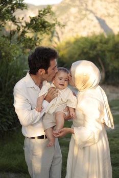 Loving parents kissing their baby in a sunny outdoor setting, capturing a warm family moment.