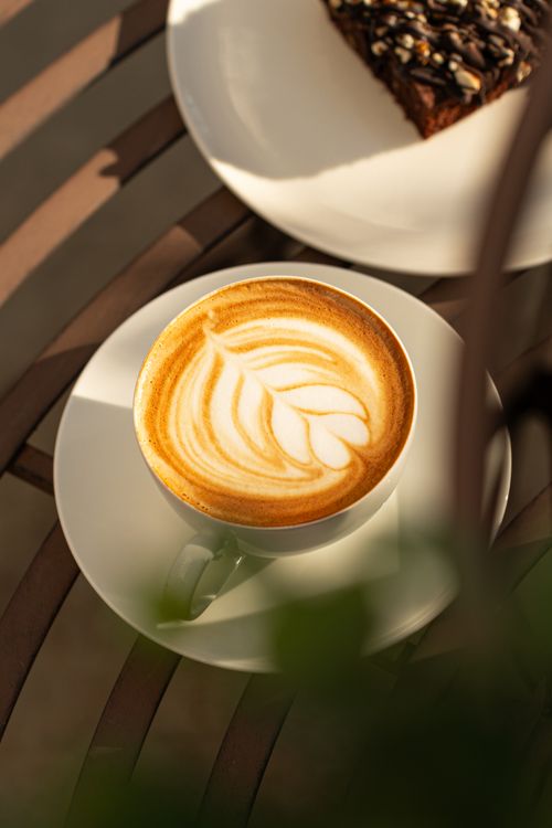 Free A cup of cappuccino with intricate latte art on a cafe table, paired with a chocolate cake slice. Stock Photo