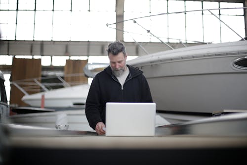 Confident adult bearded man in black clothes standing at workbench near motorboat and working with laptop in spacious hangar