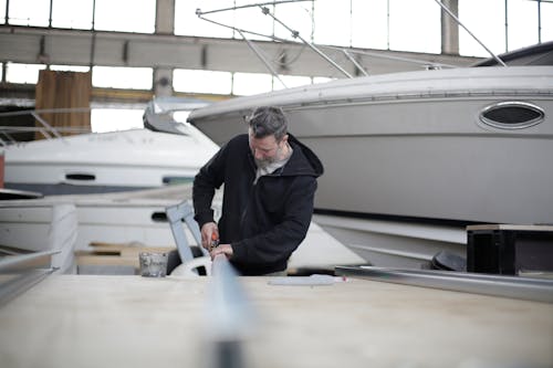 Serious adult bearded mechanic in black clothes standing at workbench and working with metal detail near motorboat in spacious hangar