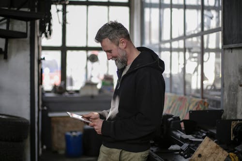 Free Side view of positive adult bearded male mechanic wearing black clothes standing near workbench and using tablet while working in spacious workshop Stock Photo