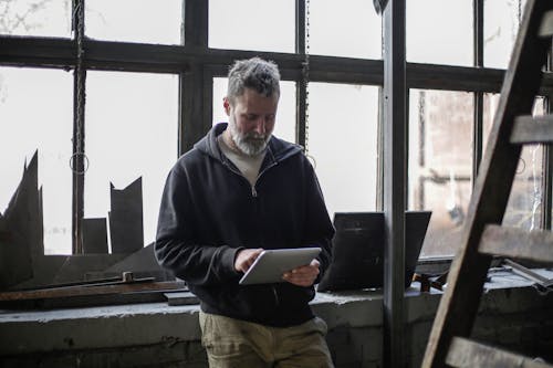Serious adult bearded workman wearing workwear standing near window and browsing tablet while working in workshop