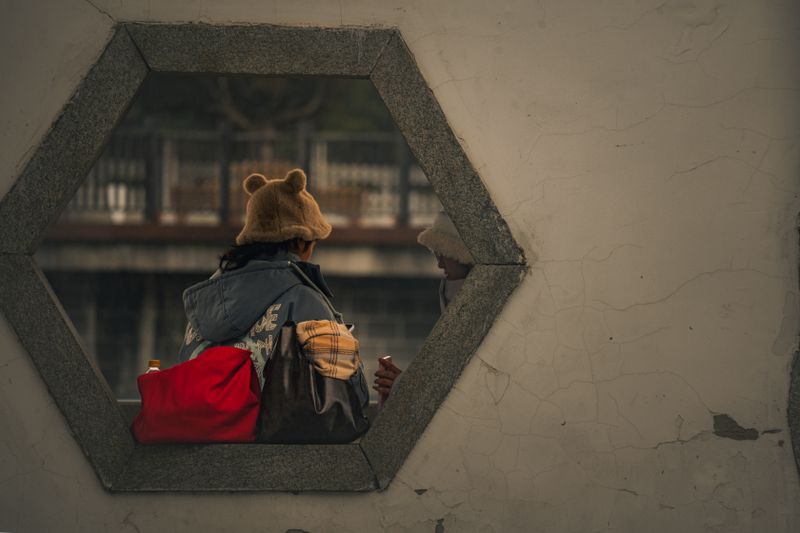 Two people in warm clothing viewed through a hexagonal stone window on a cool day.