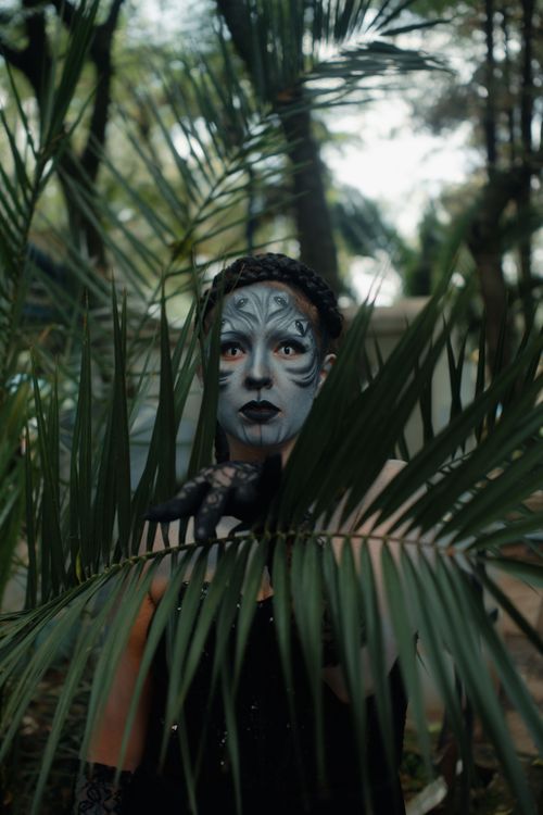 Free A person with mystical makeup peers through lush greenery, capturing a dramatic and fantasy-like scene. Stock Photo