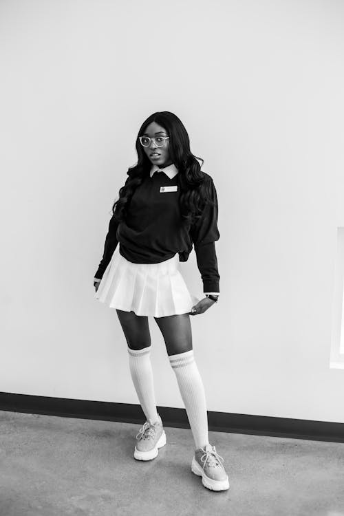 Full length of black and white African American female in trendy school uniform and eyeglasses standing near wall and looking at camera