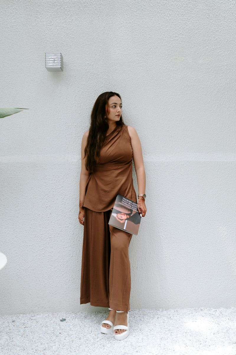 Stylish woman in brown attire poses against a minimalistic wall holding a fashion magazine