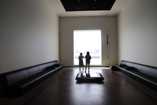 Two children stand in a minimalist gallery, looking out through a large window, creating a serene scene.