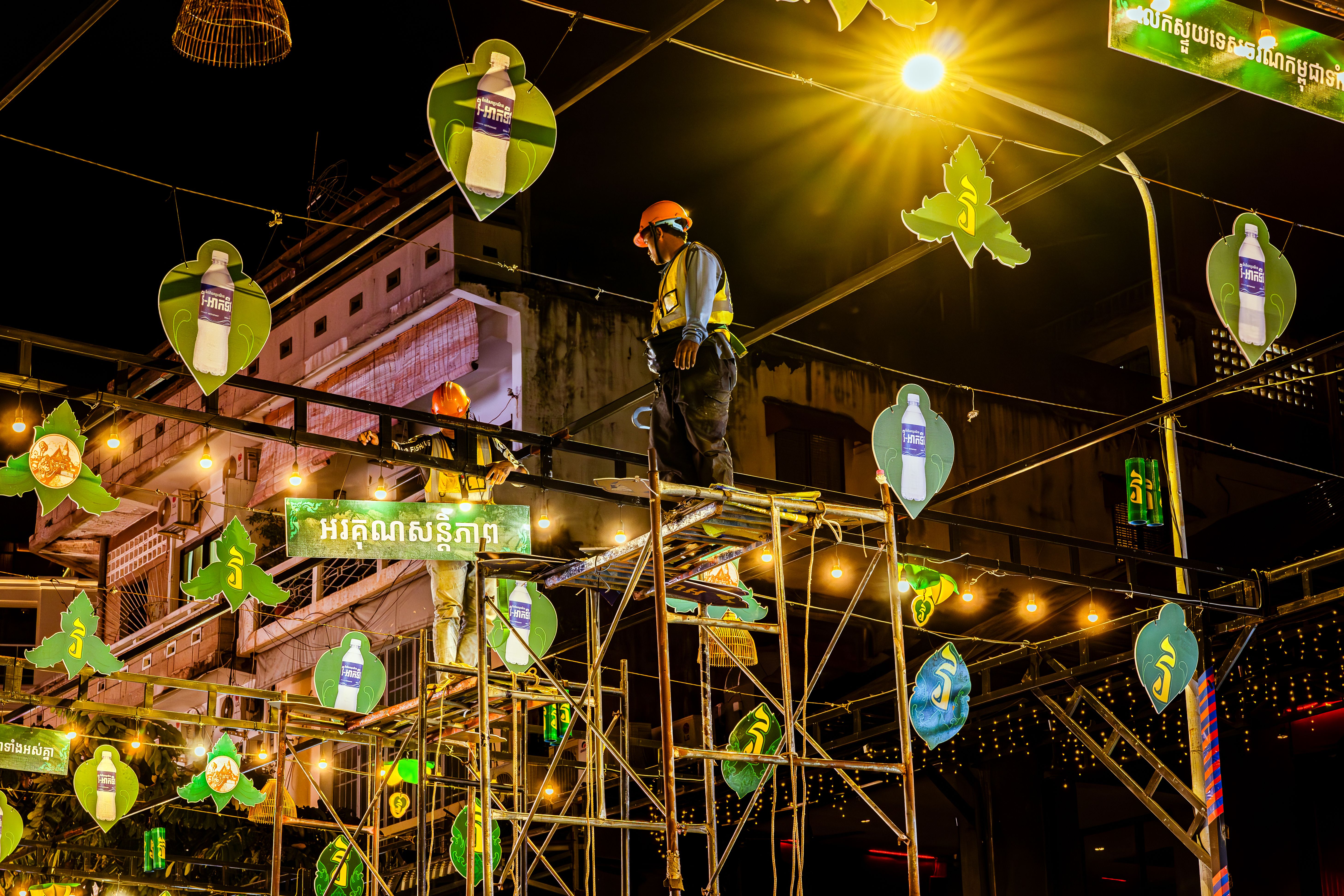 Workers installing decorative lights at night in an urban area, showcasing construction and festival preparations.