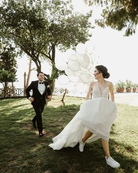 Joyful couple running in a sunny Antalya garden with balloons, capturing lively wedding vibes.