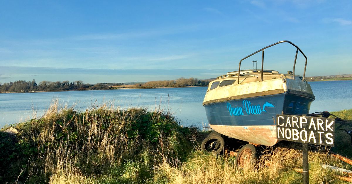 An old boat sits on the grassy shore of a serene Irish lake under a clear sky.