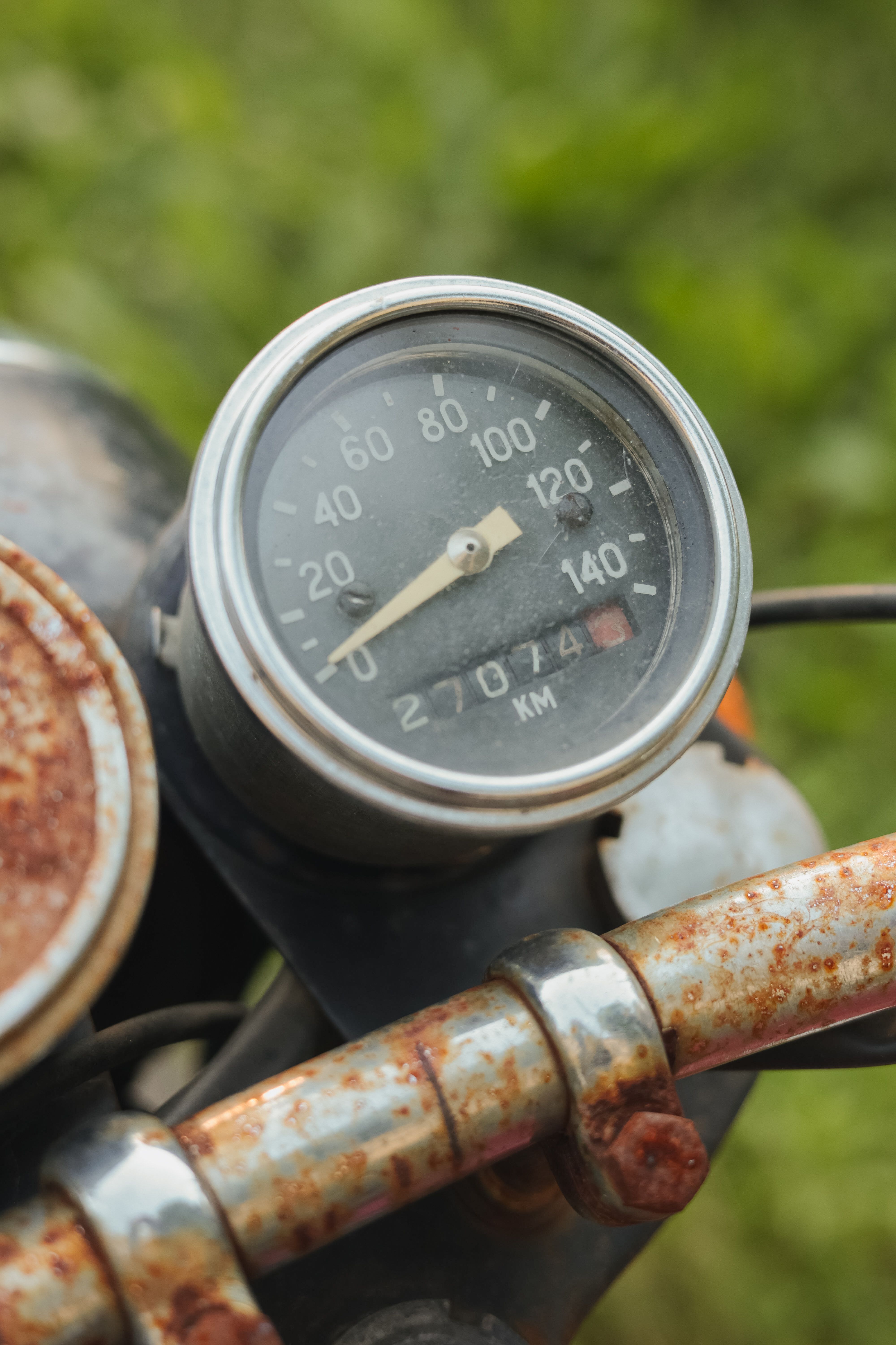 Close-up of a rusty vintage motorcycle speedometer with a background of greenery.