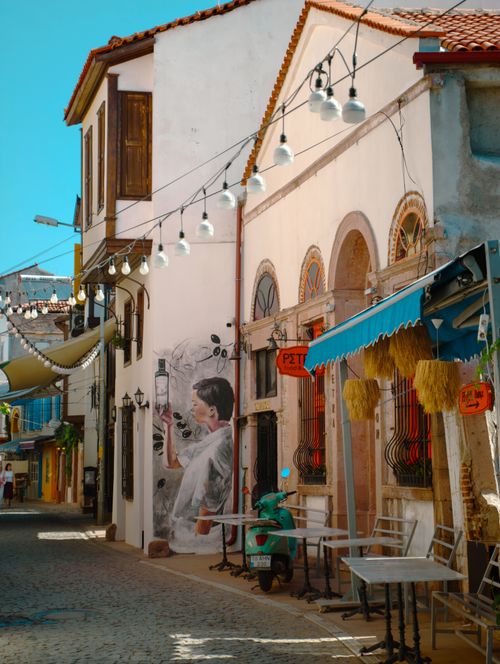 Free Quaint alley in Ayvalık, Türkiye, showcasing street art and outdoor dining in vibrant summer light. Stock Photo