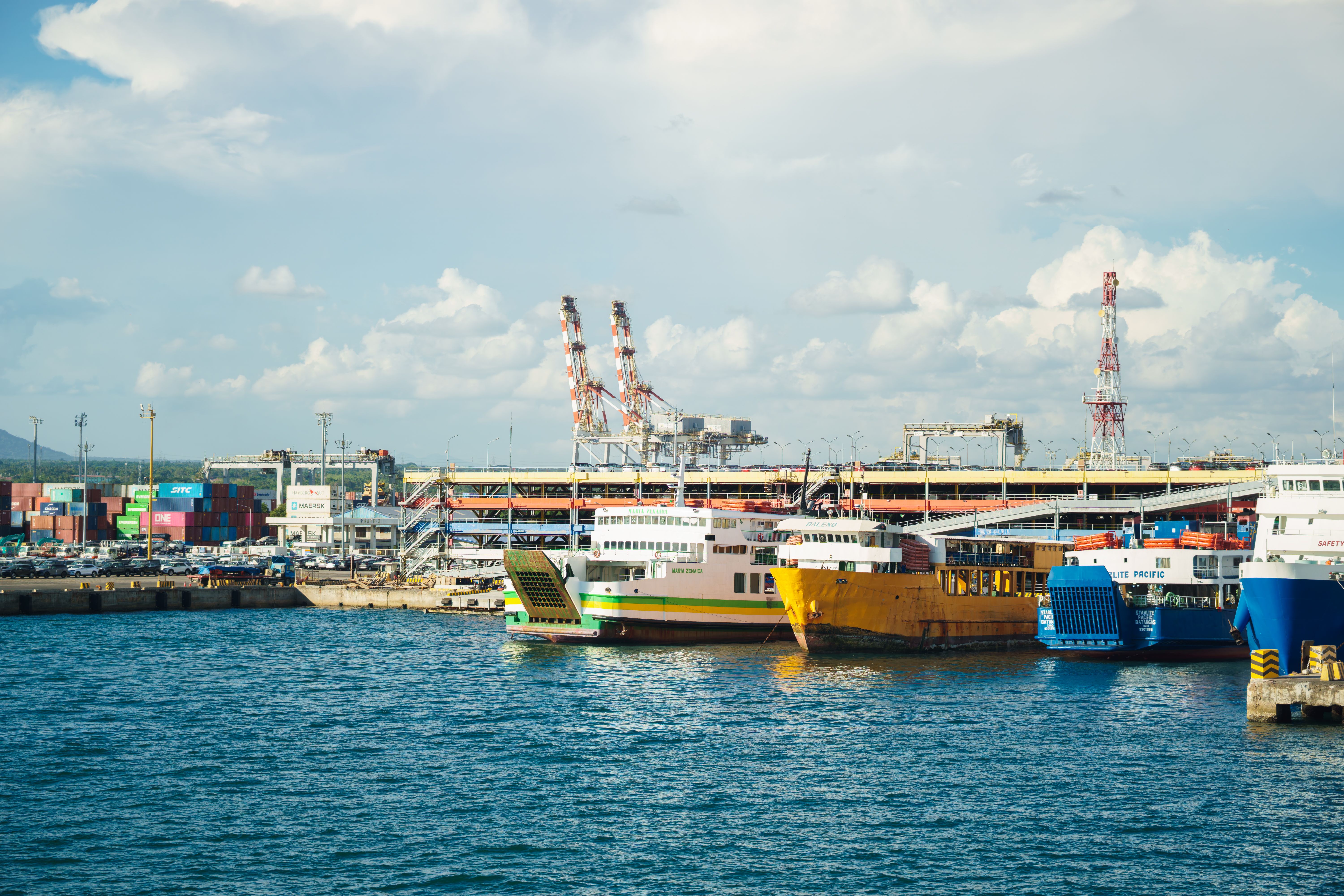 Vibrant ferries moored at an industrial port with shipping containers and cranes in the background.