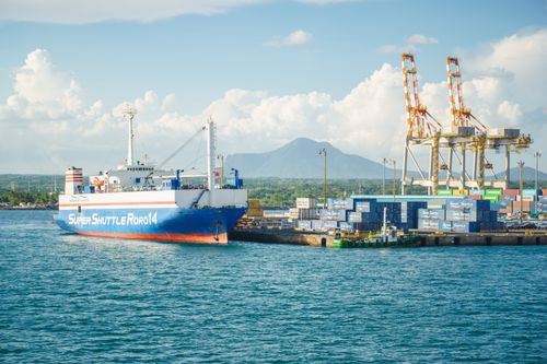 Free A cargo ship docked at an industrial port with shipping containers and cranes under a clear sky. Stock Photo