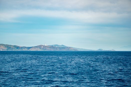 Vast ocean with distant island and lighthouse under a clear blue sky, offering a serene coastal landscape.