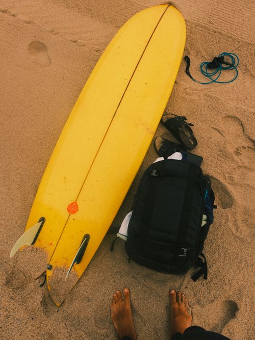 Free A yellow surfboard and backpack on a sandy beach with visible footprints, capturing a summer surf scene. Stock Photo
