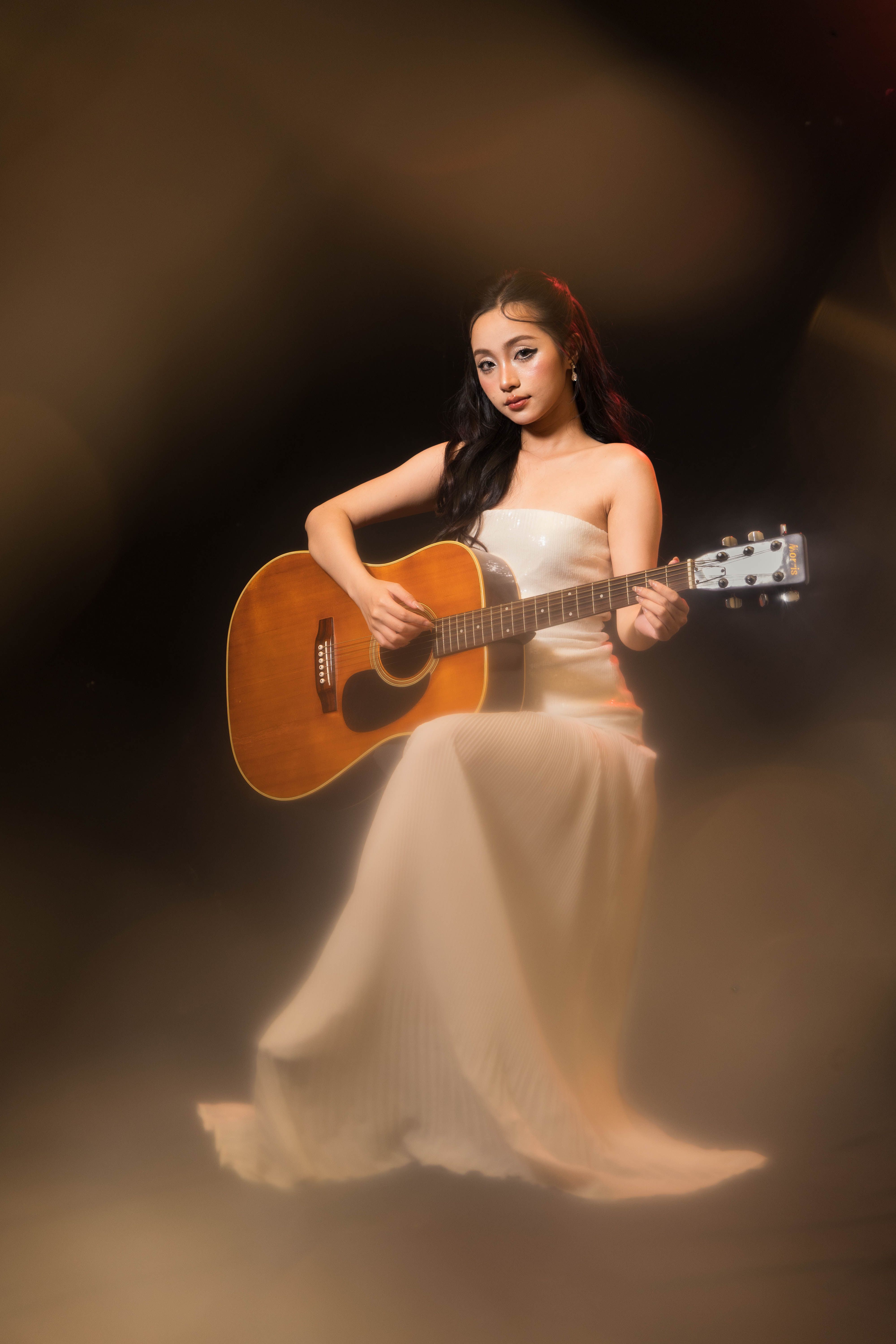 A graceful woman in an elegant dress playing an acoustic guitar under stage lighting, creating a serene and artistic mood.