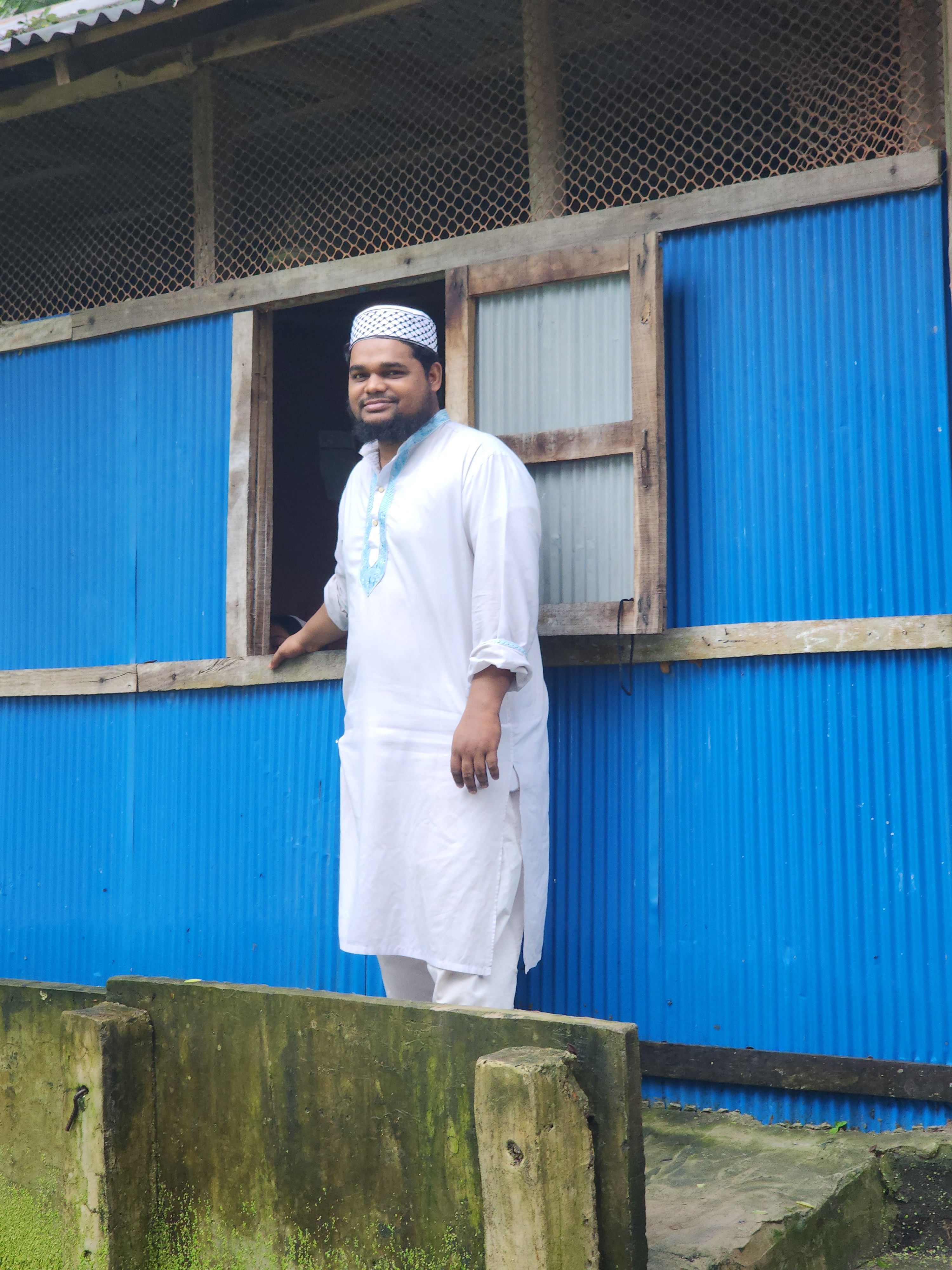 A smiling man in white traditional clothing standing by a vibrant blue building outdoors.