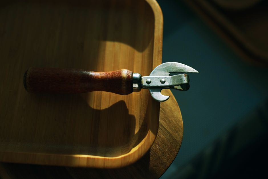 A butterfly-style manual can opener clamped onto the rim of a can, with the crank handle mid-turn and a partially cut lid visible.