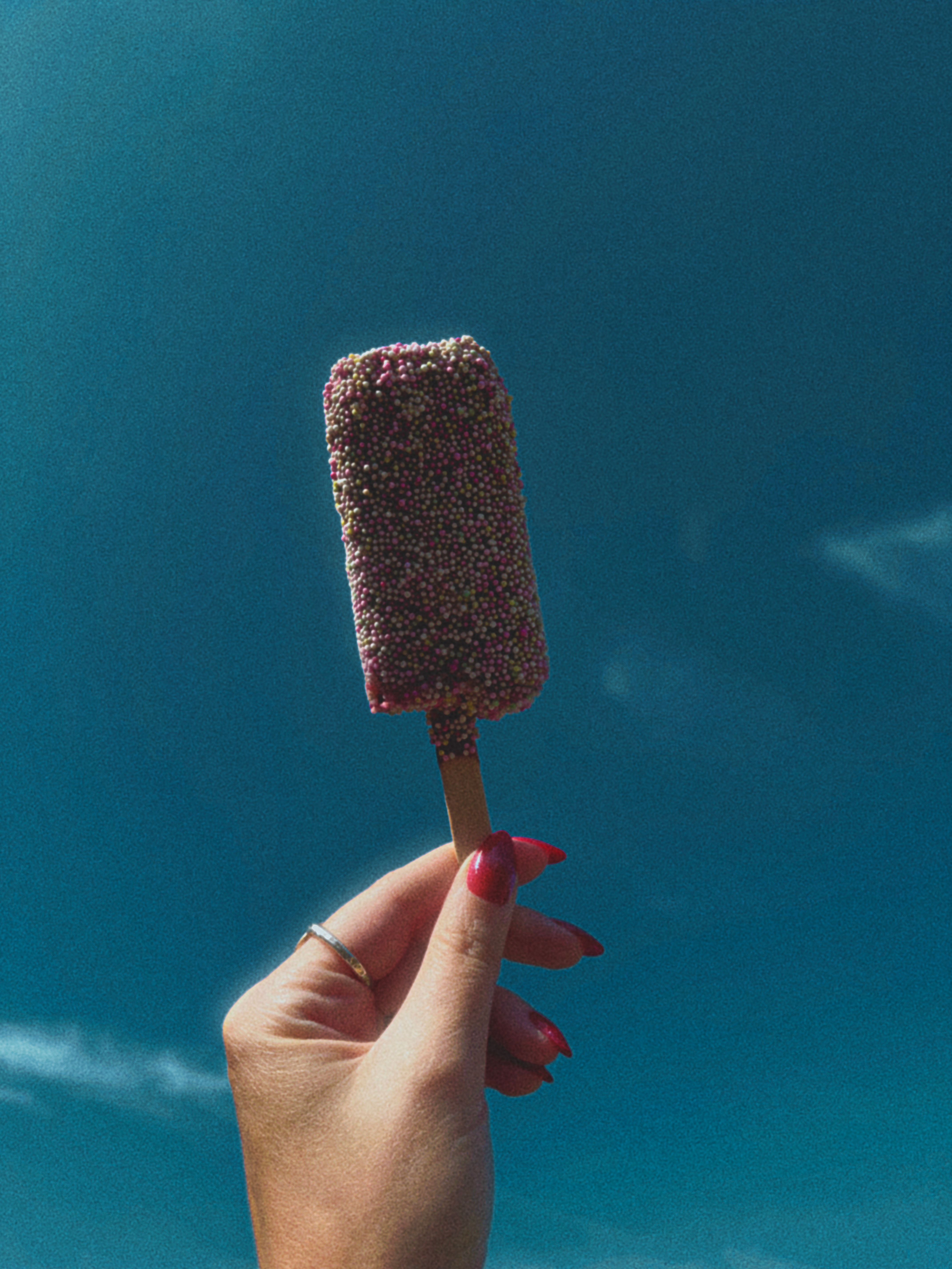 A hand holding a sprinkle-covered ice cream popsicle against a bright blue sky.