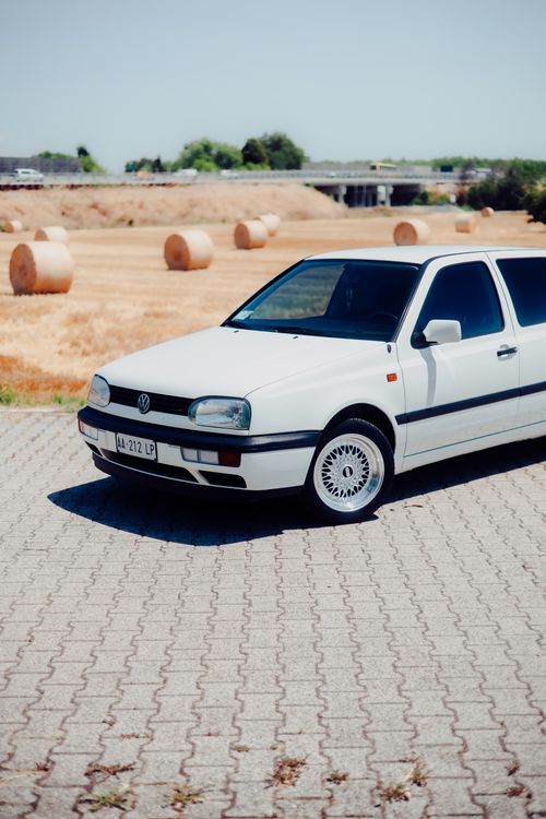 Free Classic white hatchback car parked on block pavement with a rural field backdrop in bright sunlight. Stock Photo