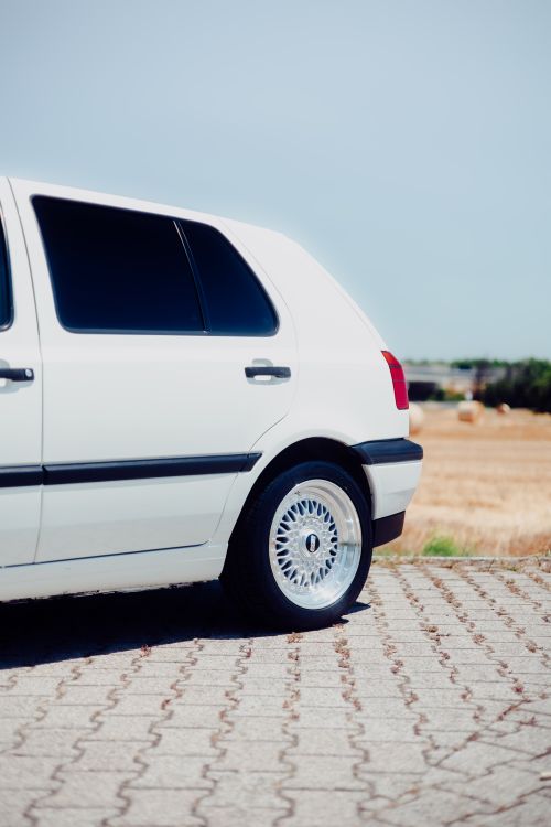 Free A white car parked on a paved road with fields in the background on a sunny day. Stock Photo