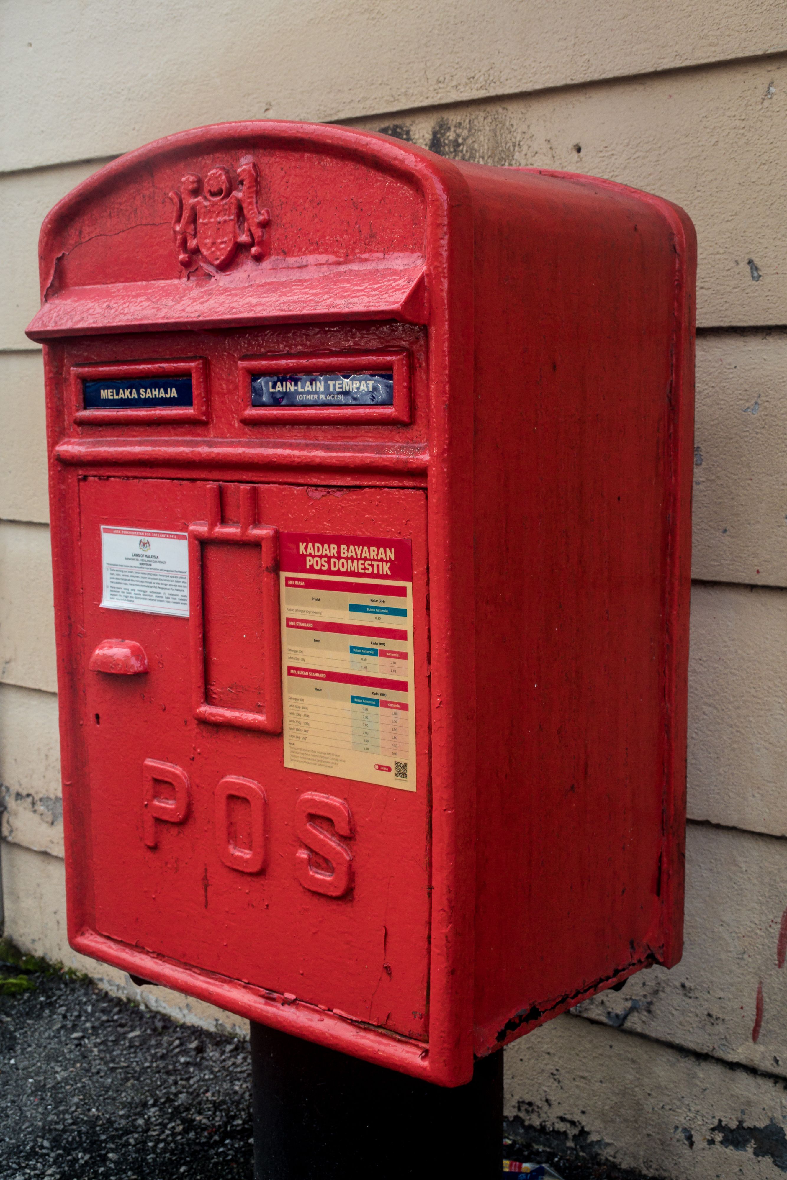 Red post box against a wall in Penang, Malaysia, showcasing local postal service details.