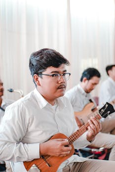 Adult musician with eyeglasses playing ukulele in a band during a music session indoors.