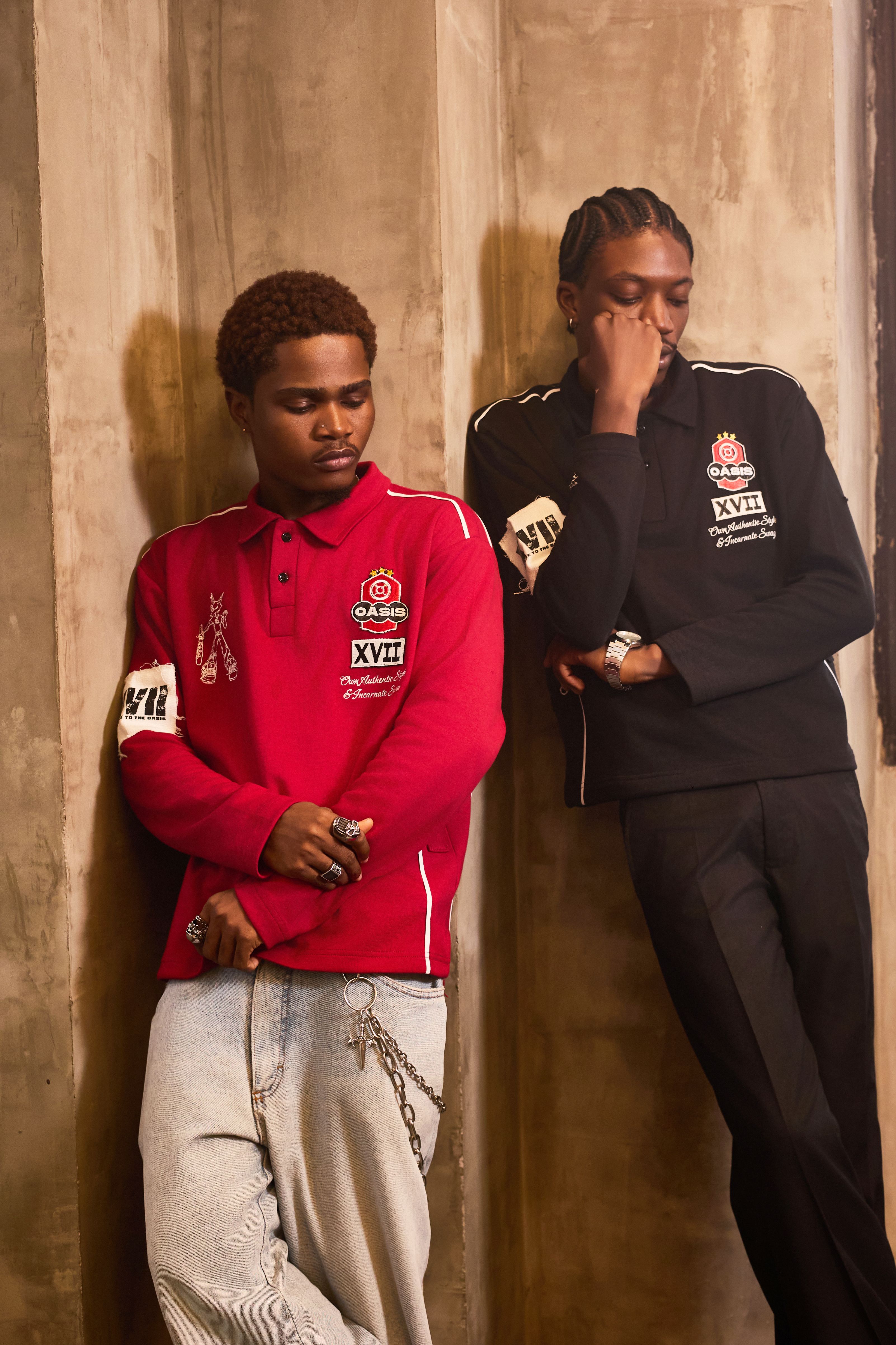 Two men in fashionable streetwear pose against a textured wall, showcasing urban style.