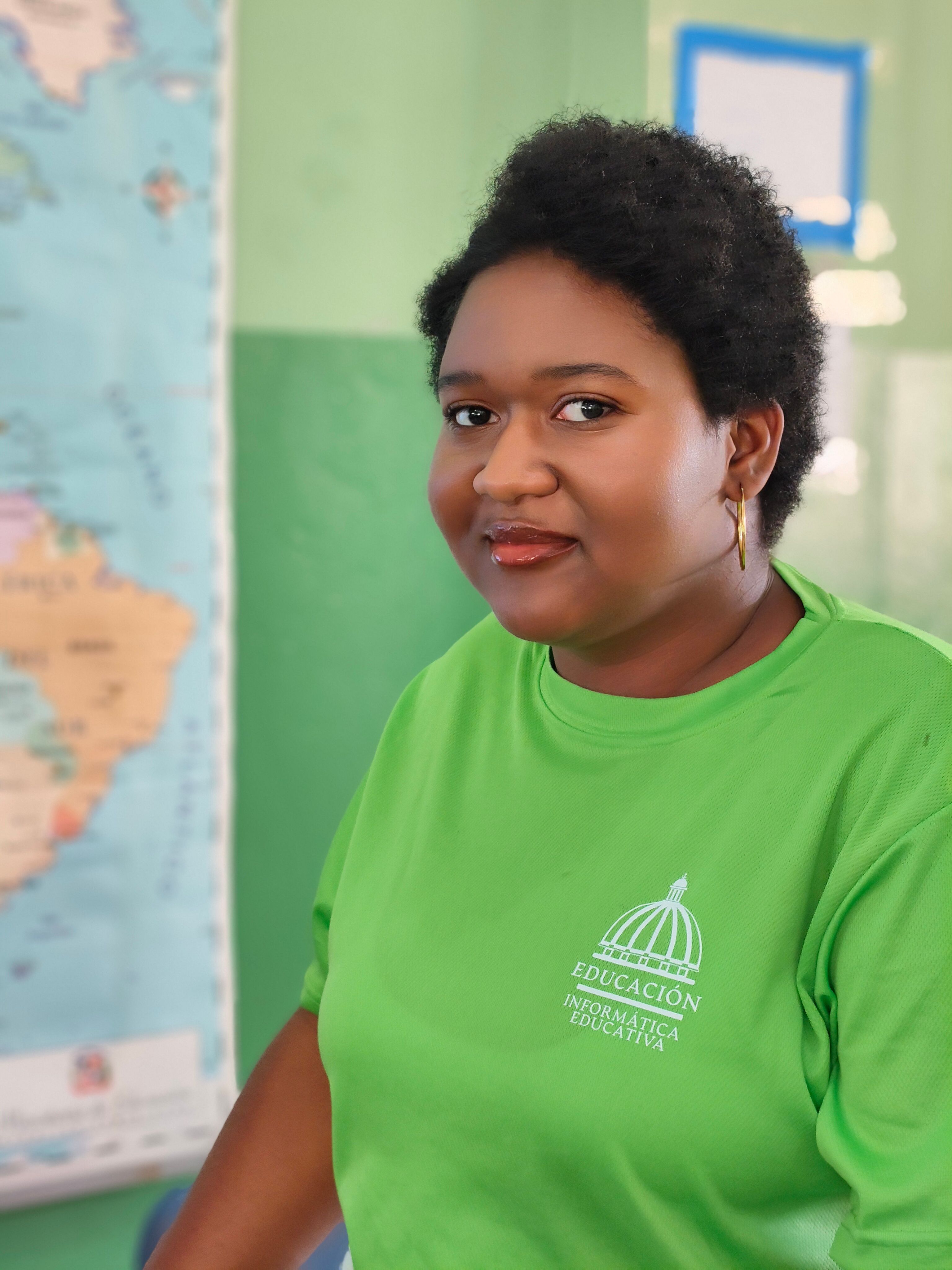 A woman in a green shirt sitting in a classroom with a map in the background.