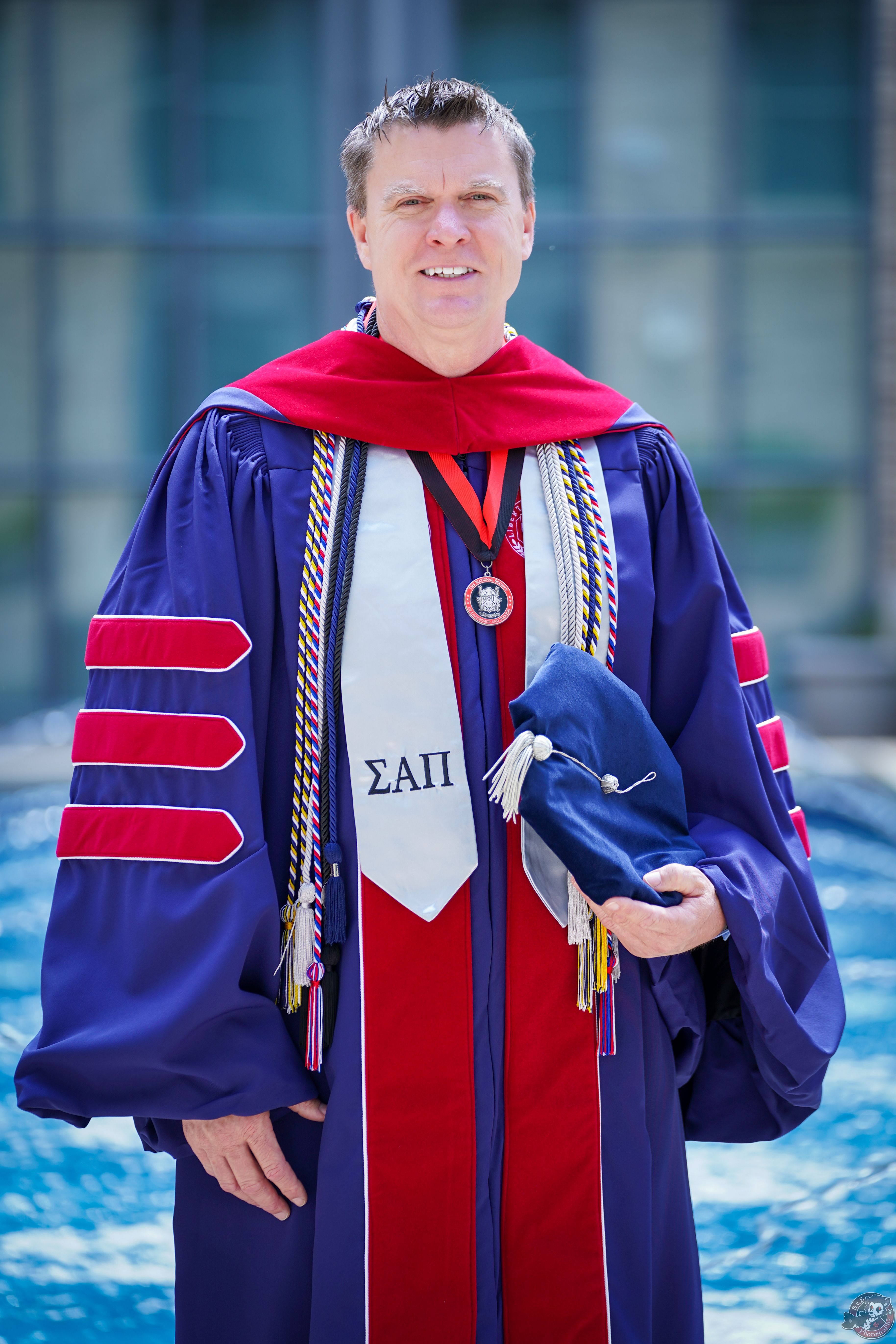 Adult male in academic regalia at an outdoor graduation ceremony.