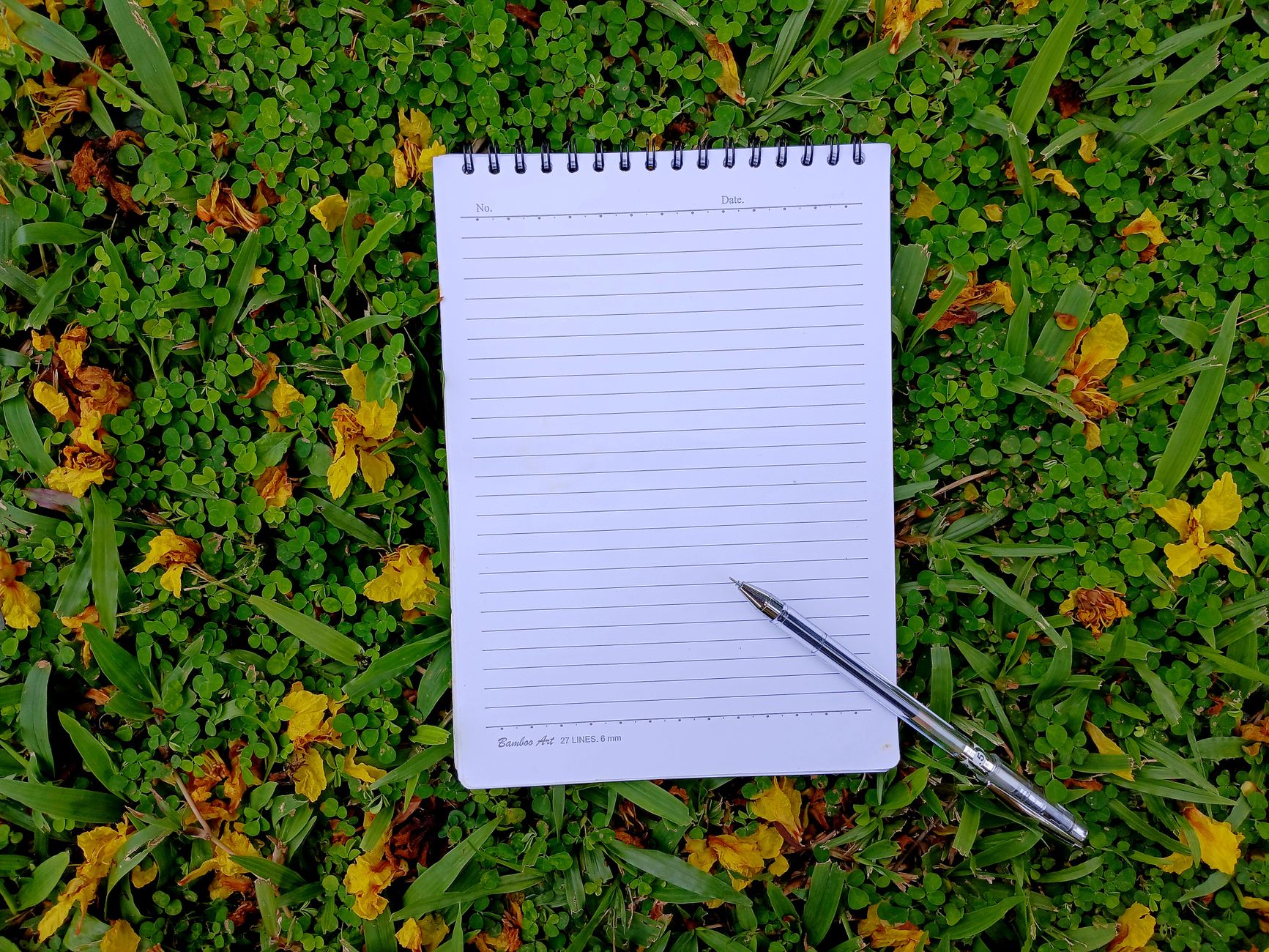 A spiral notebook and pen placed on vibrant green grass with scattered yellow flowers.