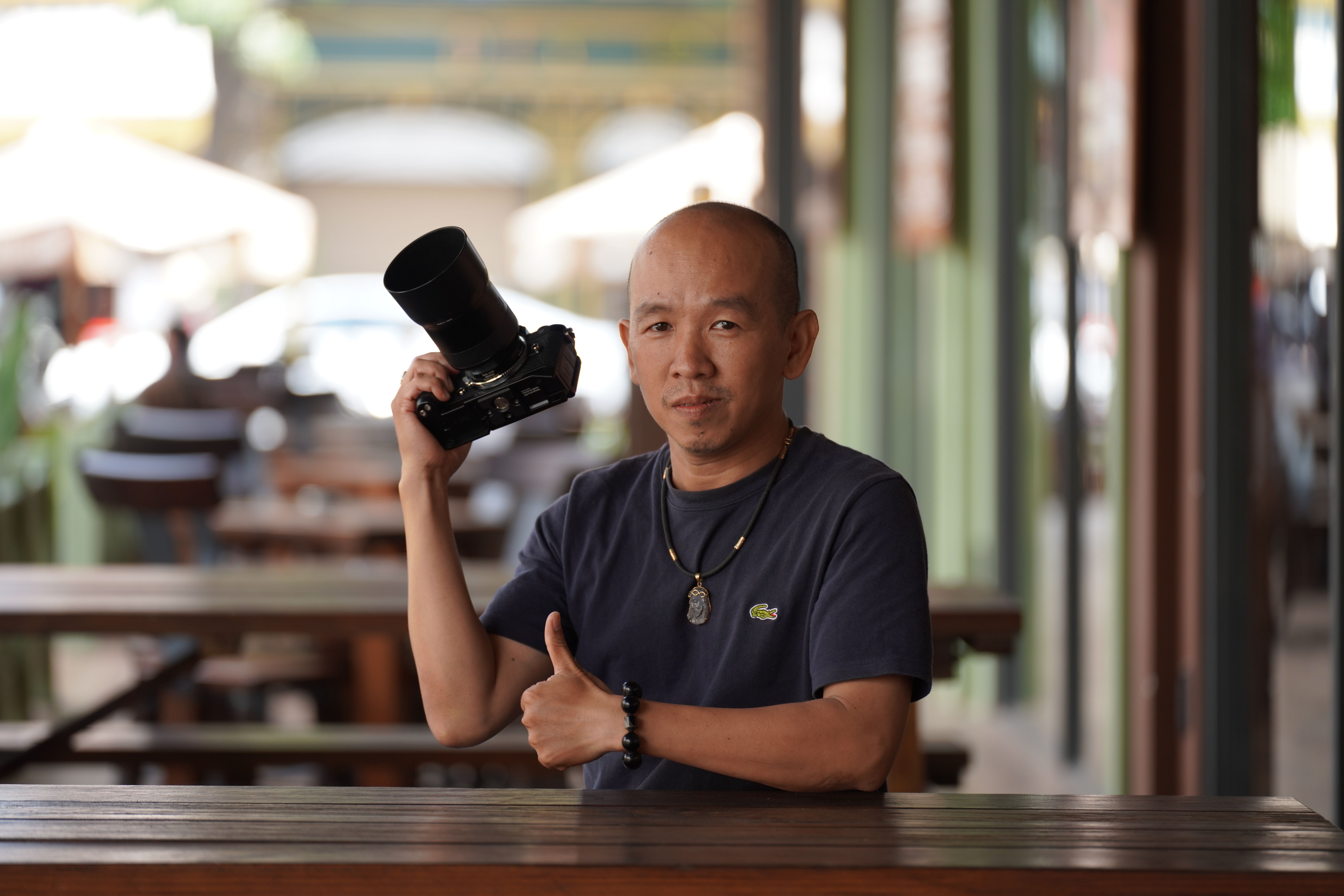 A photographer confidently holding a camera in an outdoor cafe, giving a thumbs up, suggesting enthusiasm and professionalism.