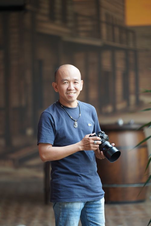 Free A smiling male photographer holding a camera, standing indoors with a blurred background. Stock Photo