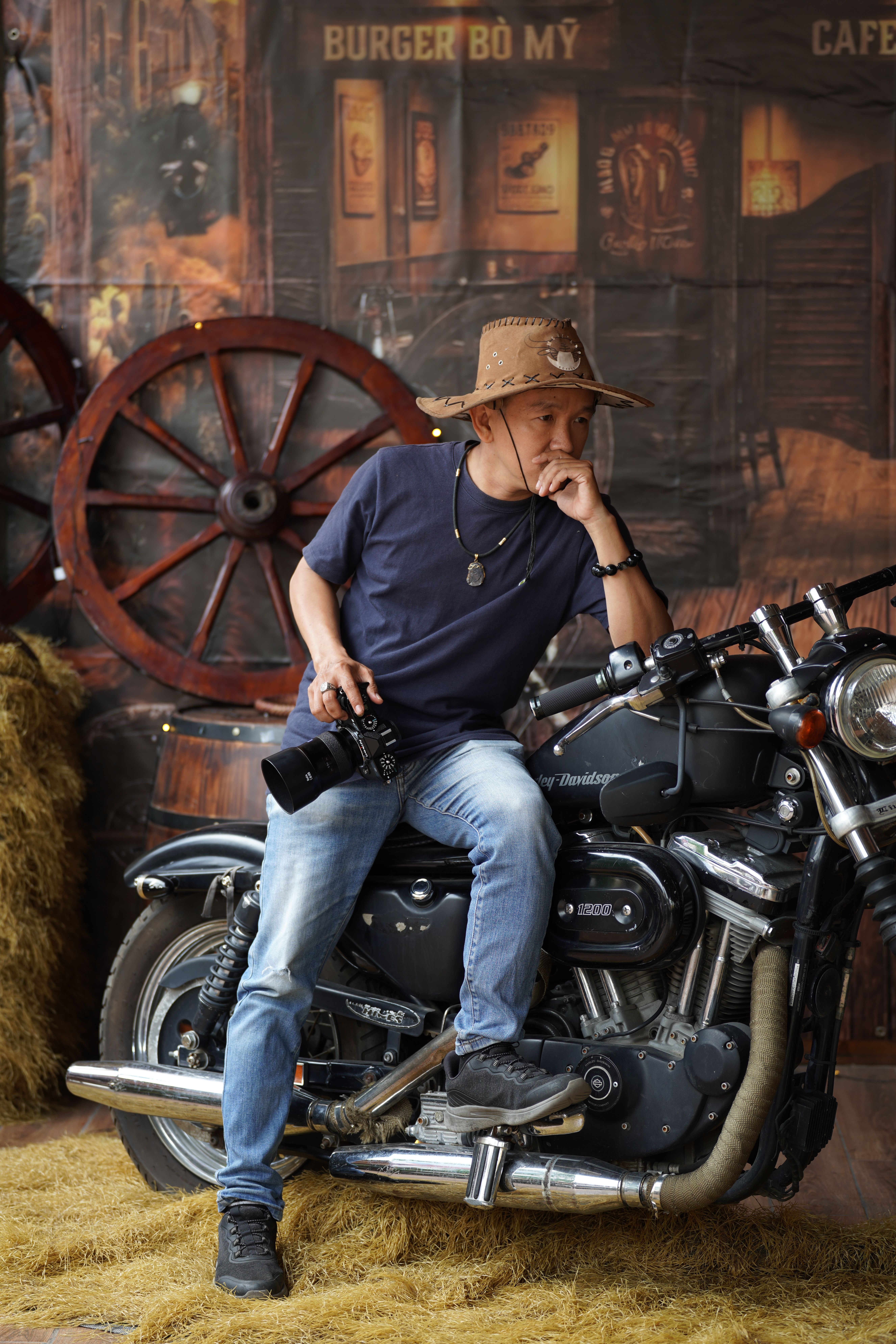 Man in straw hat holding a camera sits pensively on a Harley-Davidson motorcycle, surrounded by rustic elements.