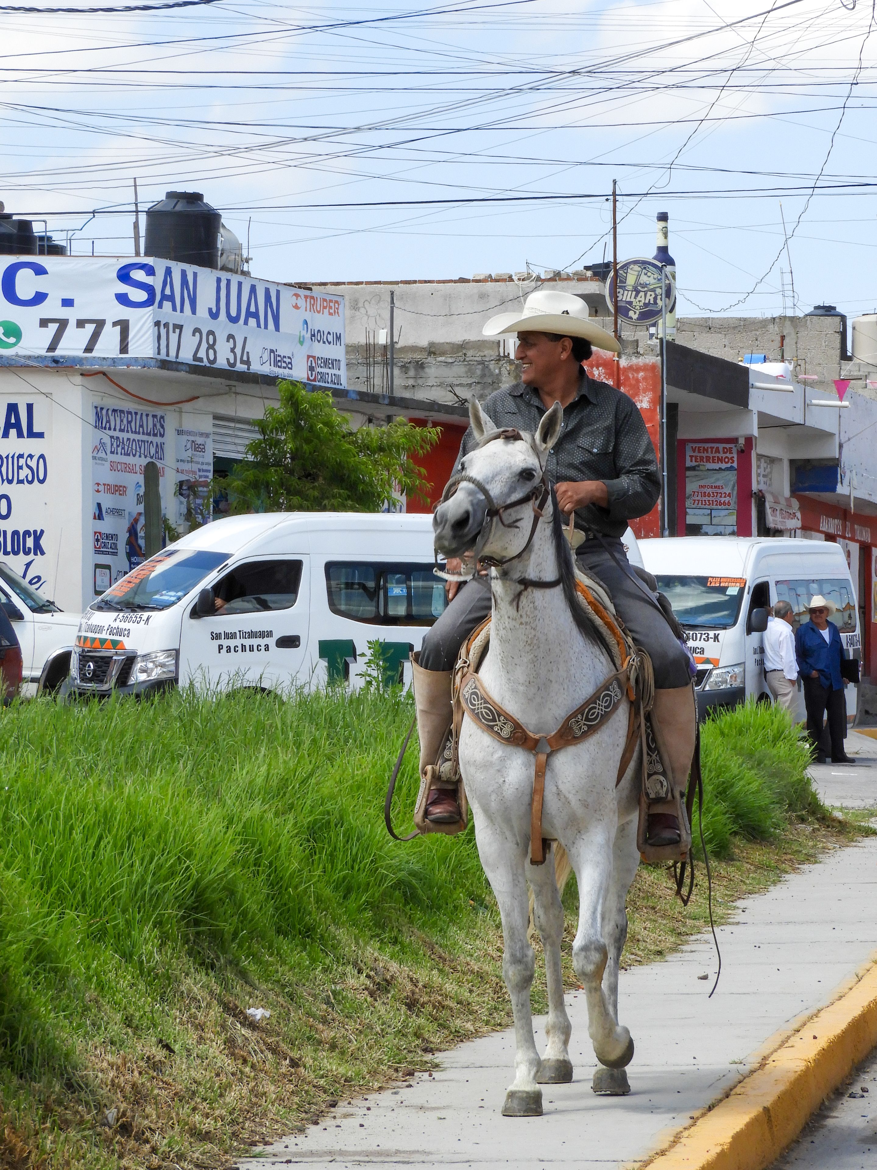 A man in traditional attire rides a white horse along a busy Mexican street, capturing a blend of tradition and modernity.