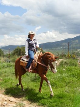 A woman rides a horse through the picturesque countryside of Mexico, with mountains and blue skies in the background.