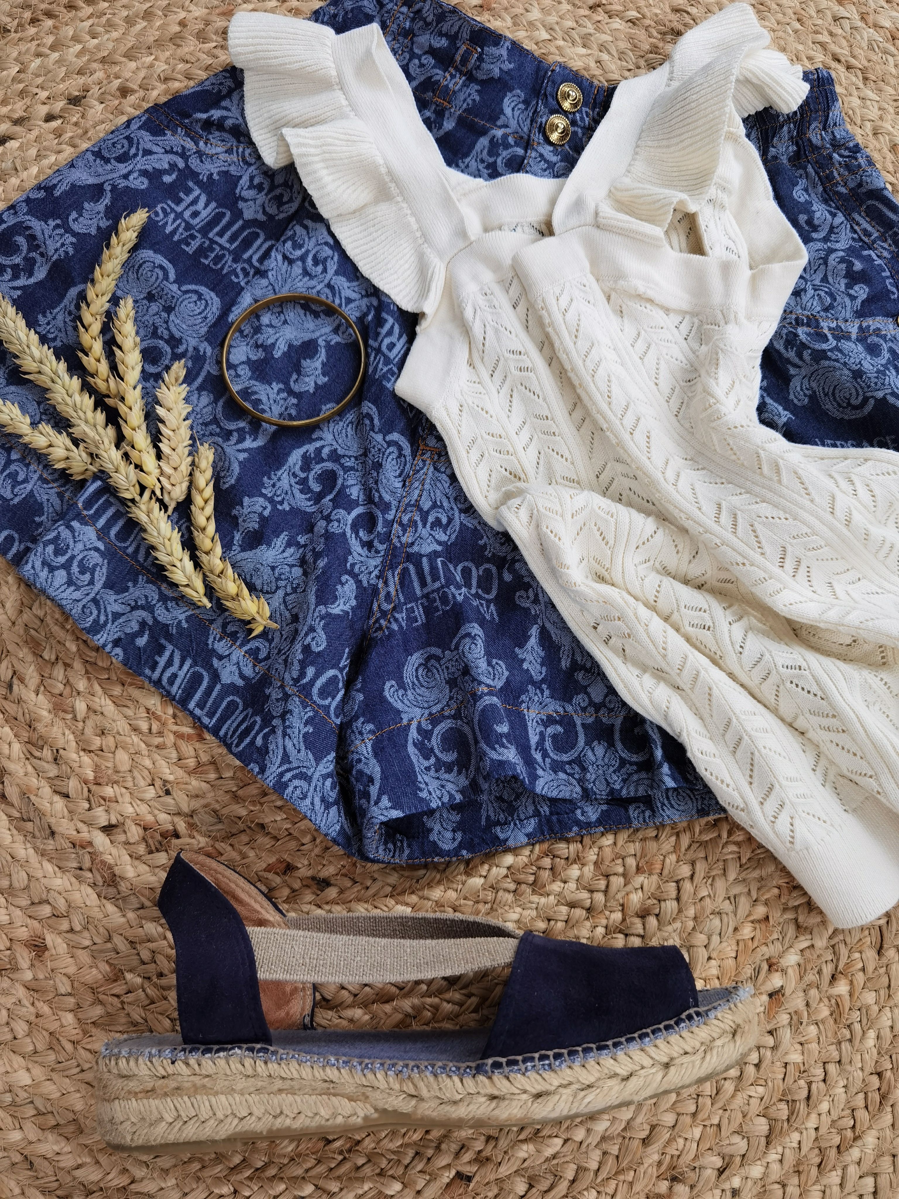 Stylish flat lay featuring blue patterned skirt, white knit top, and navy espadrille on woven mat.