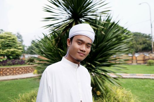Smiling adult male wearing traditional Muslim attire with a headscarf, posing outdoors in a green landscape.