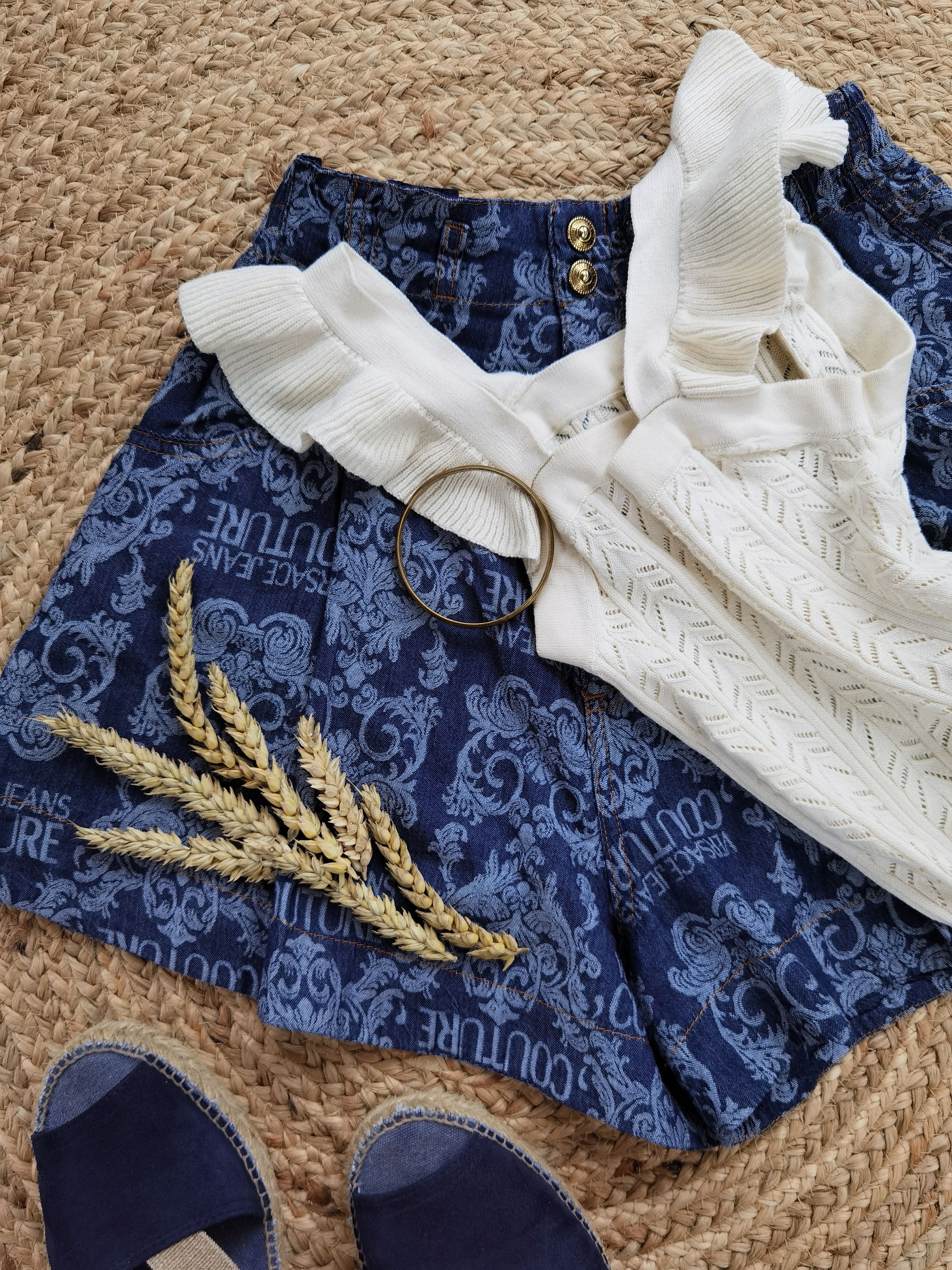 Flat lay of a chic summer outfit with blue shorts, white top, and gold accessories on a woven mat.