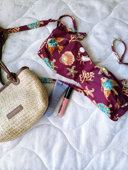 Flat lay of bikini, cream sunblock, lip gloss, and beach bag on a white quilted background.