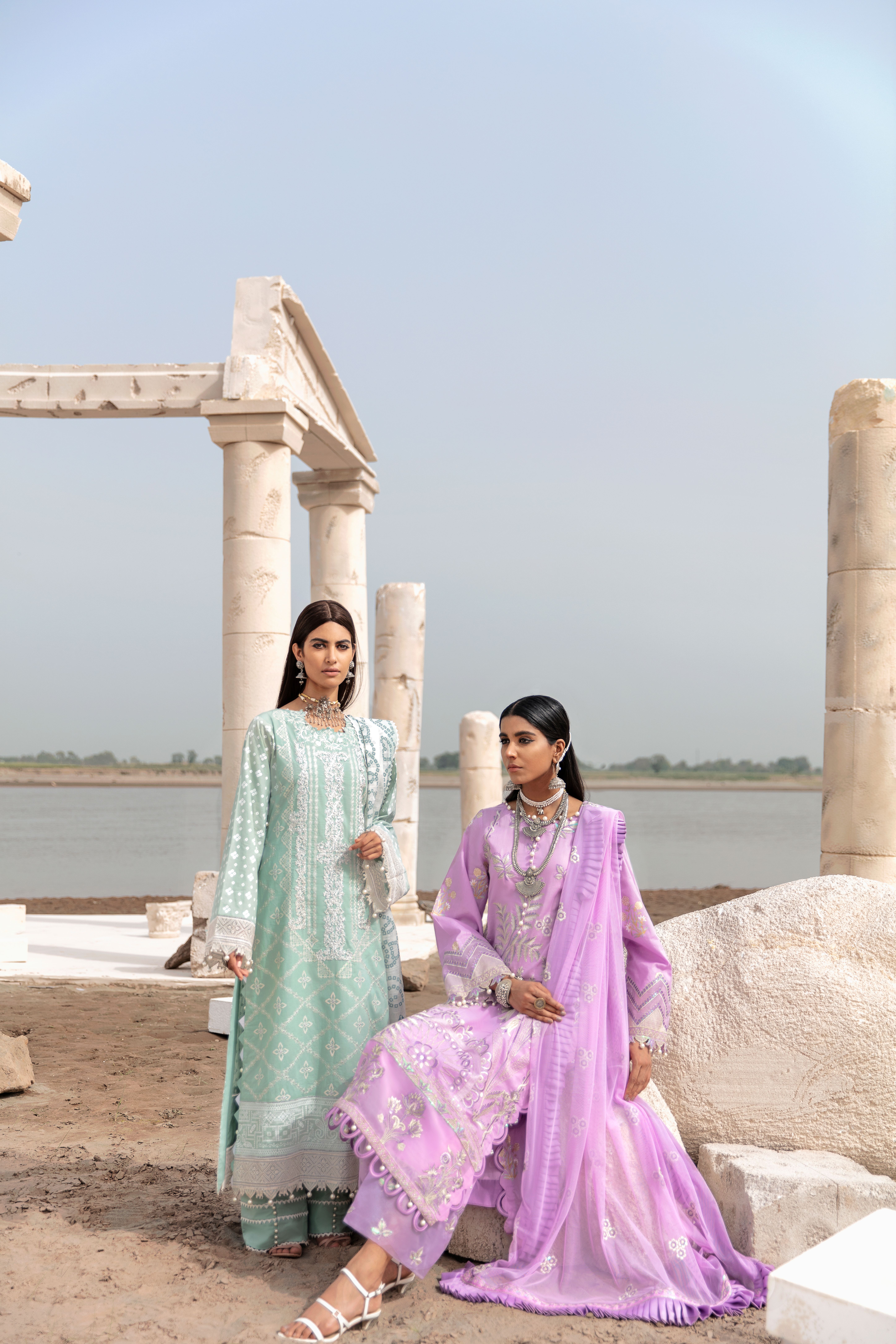 Two women in traditional attire pose elegantly near ancient ruins by a lake.