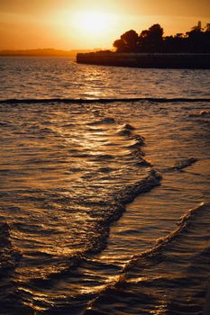 Serene sunset casting golden hues over gentle ocean waves near a silhouetted shoreline.