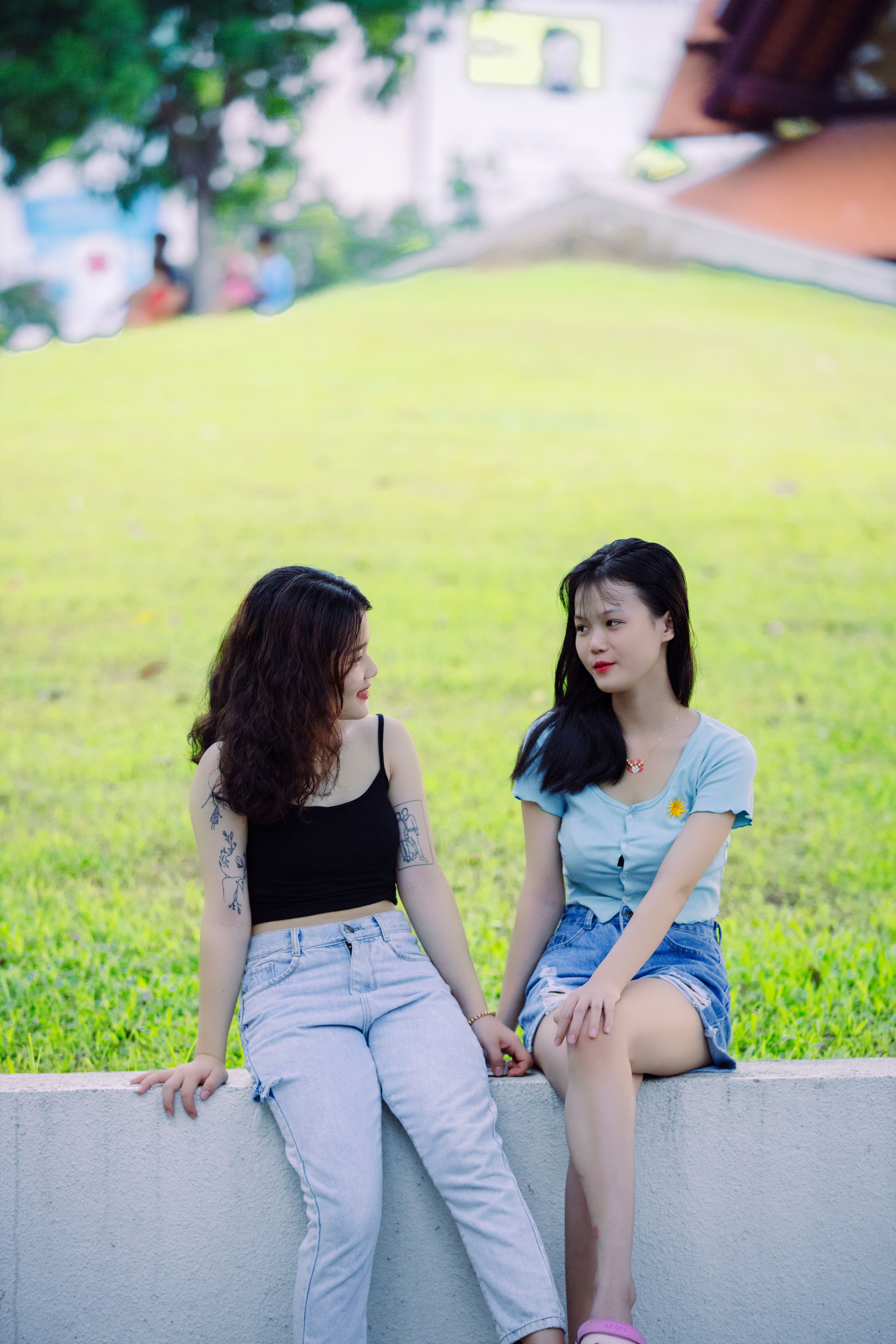 Two young women sit on a wall in a park, enjoying a sunny day together.