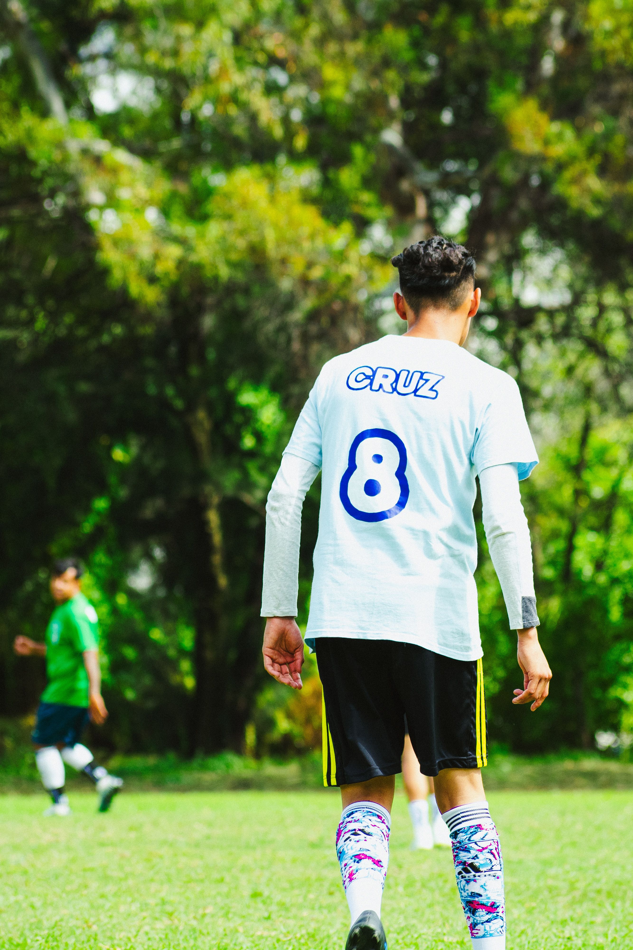 A young soccer player in a jersey stands ready on a grassy field, surrounded by greenery.