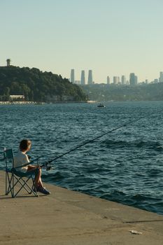 A child enjoys fishing by the Bosphorus with Istanbul skyline in the background during sunset.